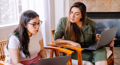 A woman and teen girl sit in armchairs indoors, discussing or working together with laptops.