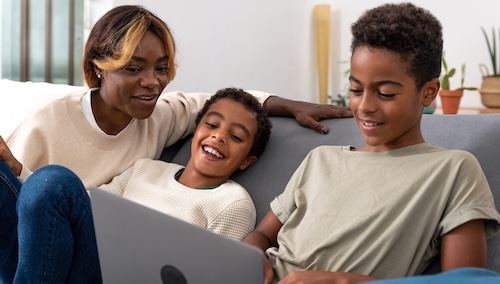 A woman and two boys smile together while looking at a laptop on a sofa.