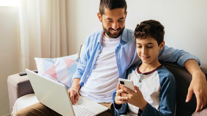 A father on his laptop sitting next to his son who is using a phone.