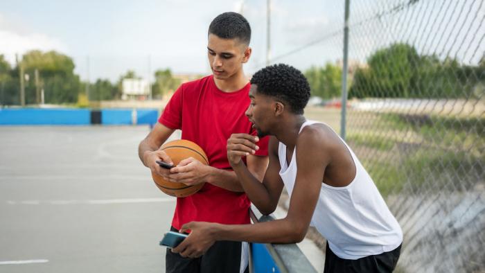 Two teen boys standing in a basketball court while holding a basketball look at their phones.