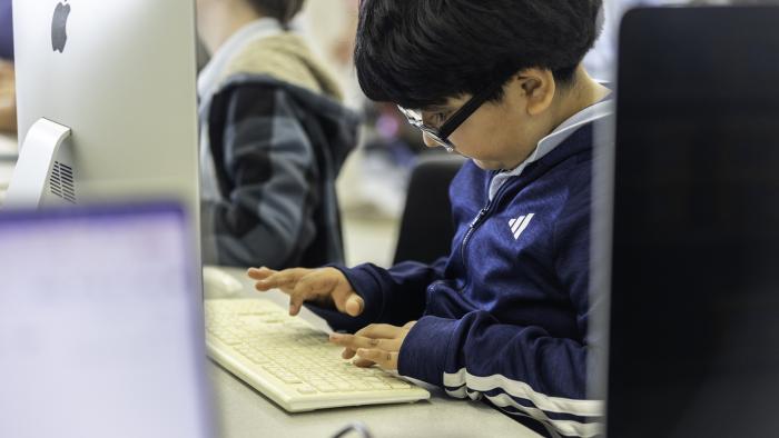 Child in a blue jacket typing on a computer keyboard in a classroom.