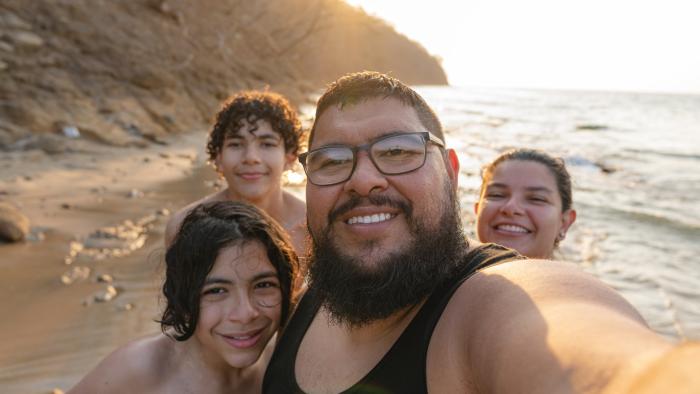 A group selfie of four people on a beach at sunset with a rocky cliff and ocean in the background.