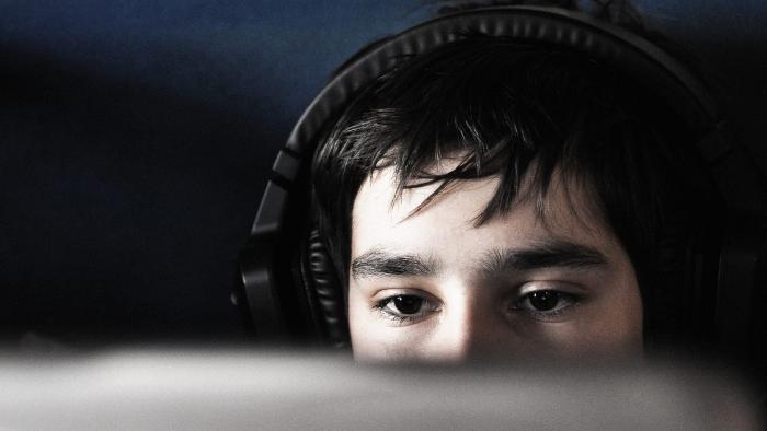 A teen boy wearing headphones looks at a computer in a dark room.