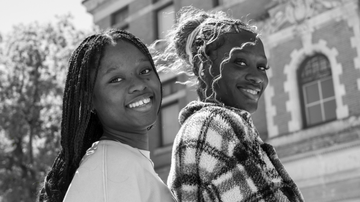 Two teen girls stand back to back outside a brick building, turning toward the camera and smiling.