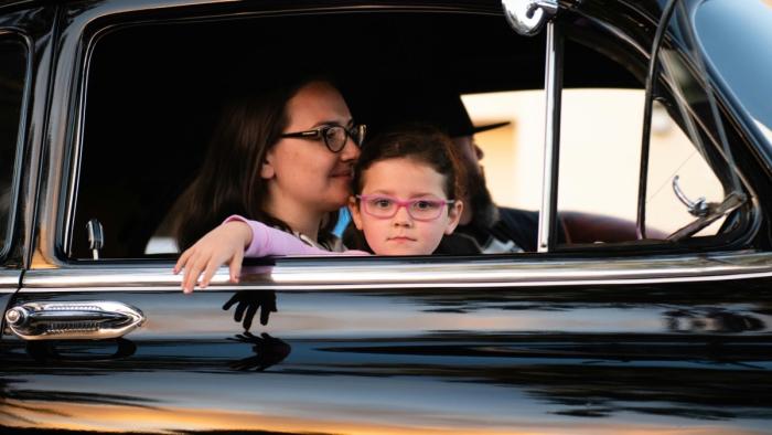 This is a photo of a mom sitting in the passenger seat of a car with a young child sitting on her lap. The mom is looking forward while the child look
