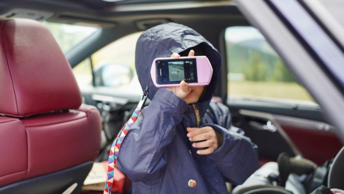 This is an image of a child in the backseat of a car. They're holding a smartphone in front of their face and on the screen is an image of a cute dog.