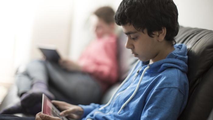 Teen boy wearing a blue sweatshirt uses his tablet on a couch.