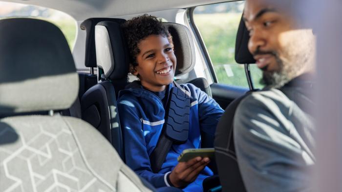 A smiling child in a blue jacket holds a smartphone in the backseat of a car with a man in the front seat.