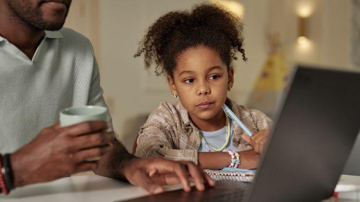 A young girl with curly hair looks at a laptop, sitting beside an adult holding a mug.