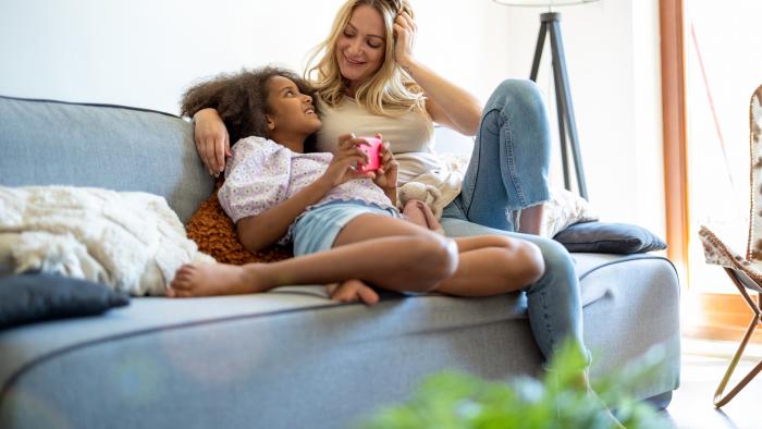 A woman and child holding a device sitting on a gray couch, smiling at each other.