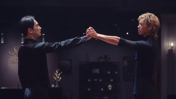 10Dance movie: In a dark dance studio, Japanese man with short black hair, left, reaches out and clasps hands with Japanese man with blond hair, right