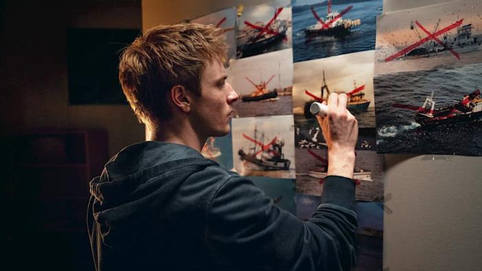 23 000 Lives movie: Louis Hoffman stands at a board filled with pictures of ships, holding a red marker