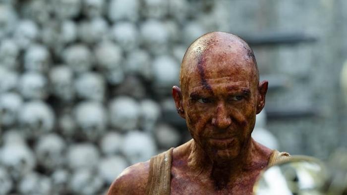 28 Years Later Movie: Dr. Kelson (Ralph Fiennes), covered in red iodine, stands in front of an out-of-focus tower of skulls