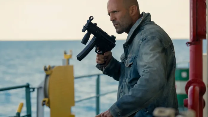 Mutiny: Shaved-headed man in denim jacket holding a gun on a boat, ocean in the background
