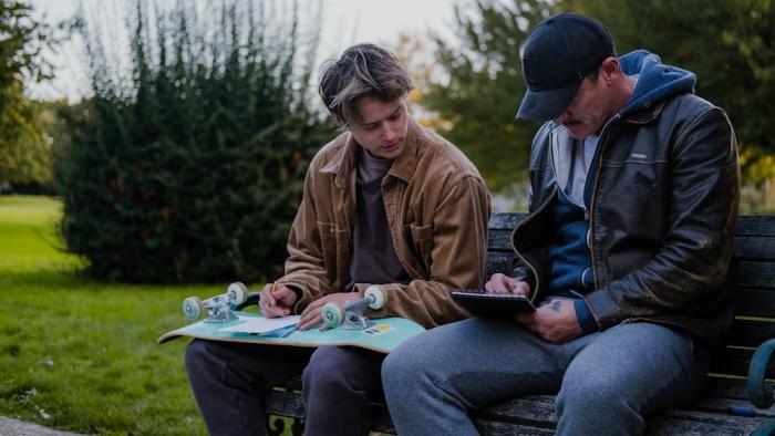 5lbs of Pressure Movie: Jimmy (Rudy Pankow) holds his upside-down skateboard on his lap, taking a drawing lesson from Adam (Luke Evans)