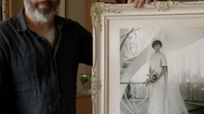 A Tree of Life movie: Anthony Fienberg, wearing a dark shirt and pants,  holds a framed picture of his mother Joyce who was killed at the Tree of Life synagogue