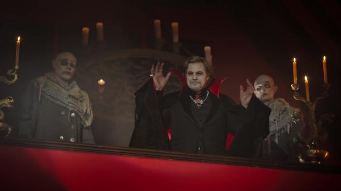 A Vampire in the Family movie: On red balcony two hairless vampires flank Brazilian Dracula in black cape as he addresses people below