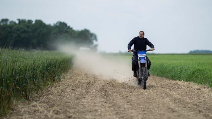 Ad Vitam movie: White French man in dark blue hoodie center right rides motorcycle through dirt path, tall grass on either side of him