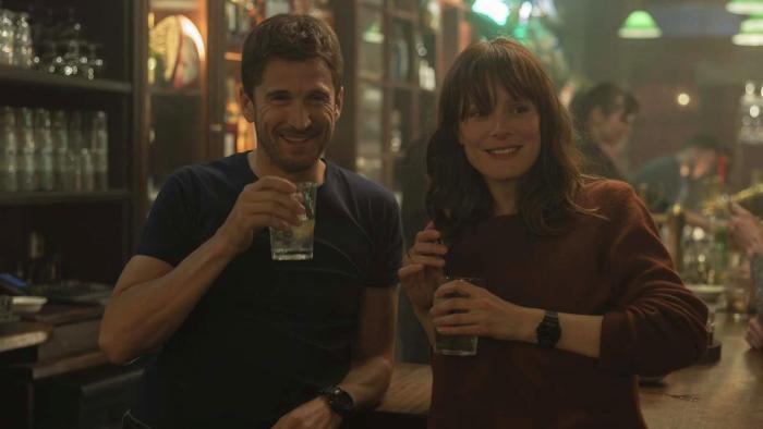 Ad Vitam movie: White French man left and White French woman right stand together smiling with drinks in hands in crowded bar