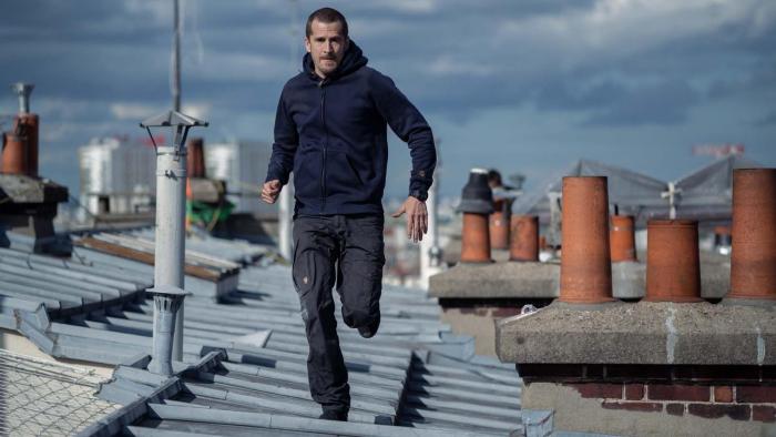 Ad Vitam movie: White French man in dark blue hoodie and black pants center runs across rooftops in Paris, France