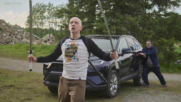 Adulthood movie: Anthony Carrigan is wild-eyed holding a sword in each hand, Josh Gad behind him with a knife