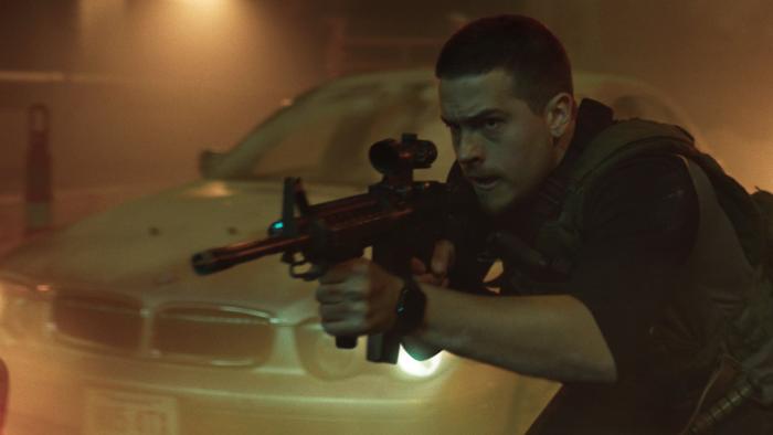 Aftermath movie: Dylan Sprouse with buzz cut hair center right holds machine ready to shoot while crouch walking next to a car