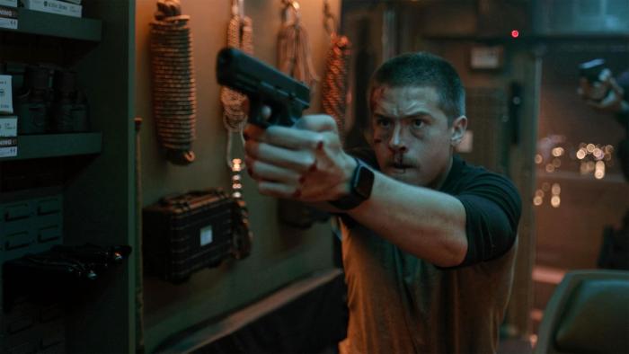 Aftermath movie: Dylan Sprouse with buzz cut hair center points a handgun at someone inside a van with ropes hanging on its wall