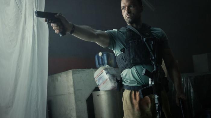 AKA: French White man in black bullet proof vest and automatic rifle hanging from neck points handgun with right hand toward left
