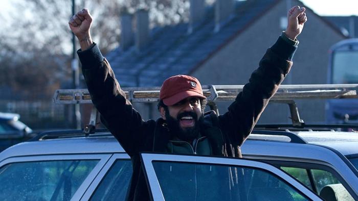 Ali & Ava: Adeel Akhtar stands in front of his car with his arms in the air, smiling.