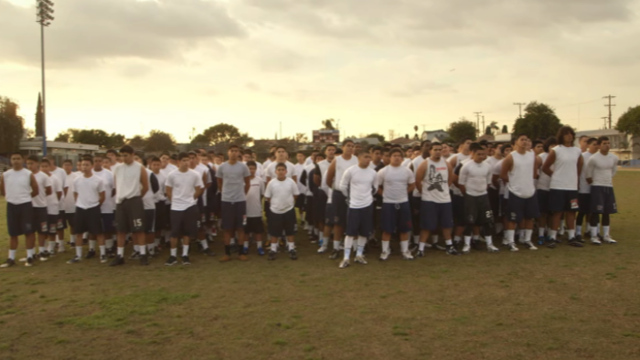 The All-Americans Movie: Scene 1 A group of football players in black shorts and white T-shirts stand together on a field. 
