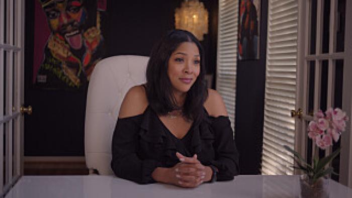 Still from All Up in the Biz: Tara Hall, the wife of Biz Markie, with a graphic illustration of her husband in the background, seated with hands folded together while in a white chair in front of a white desk, wearing a black V-neck halter top with cutout shoulders and long sleeves