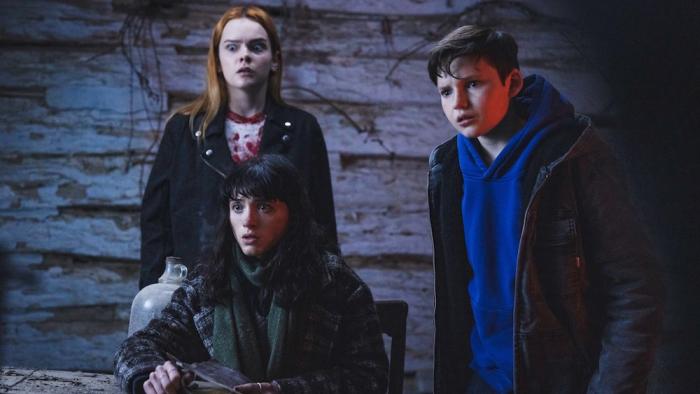 All Fun and Games Movie: In an old cabin, Billie (Natalia Dyer), who's seated; Sophie (Laurel Marsden), who's behind her; and Jo (Benjamin Evan Ainsworth), who's to the right, stare in horror at something spooky
