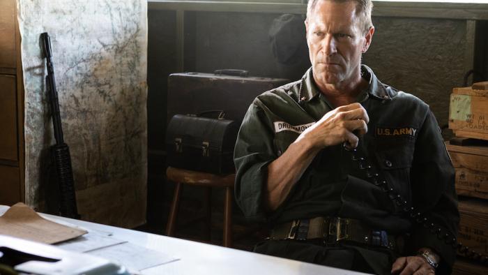 Ambush Movie: A grim-looking General Drummond (Aaron Eckhart) sits at a desk, clutching a radio handset, and a rifle leans against the wall to his left