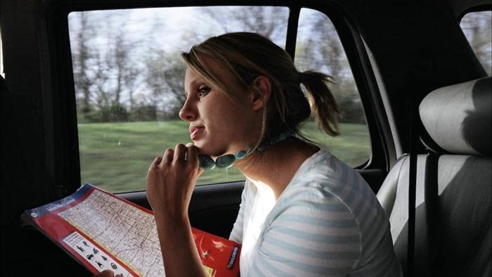 Megan Krizmanich sits in the back of a car holding a map. She is wearing a chunky necklace that she holds between her fingers.