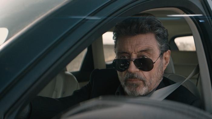 American Star Movie: Looking serious, Wilson (Ian McShane) sits in the driver's seat of his car, dark glasses pulled slightly down over his nose