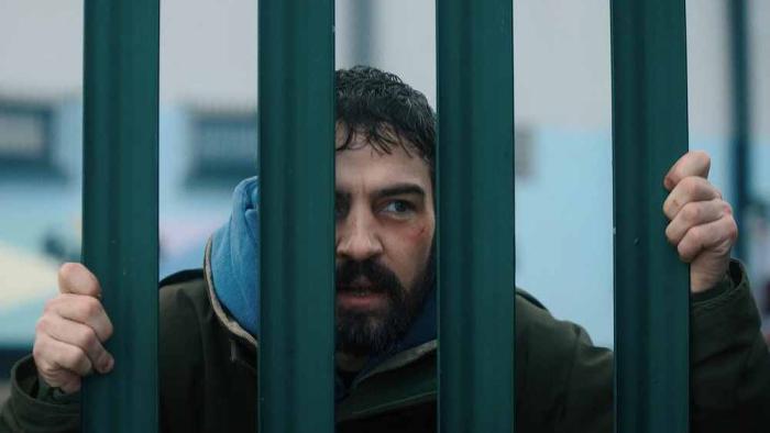 Amongst the Wolves: A bearded middle-aged White man looks through a fence
