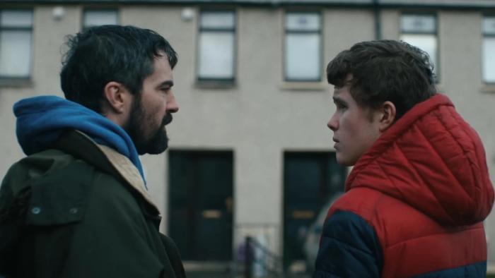Amongst the Wolves: A bearded middle-aged White man talks to a munch younger White man outside a building 