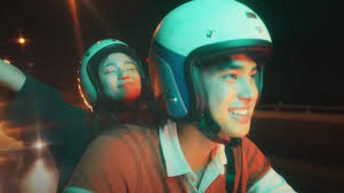 An Inconvenient Love: Filipino woman in white motorbike helmet left has arms up as she rides on back of motorcycle driven by Filipino man in orange shirt and identical white helmet center