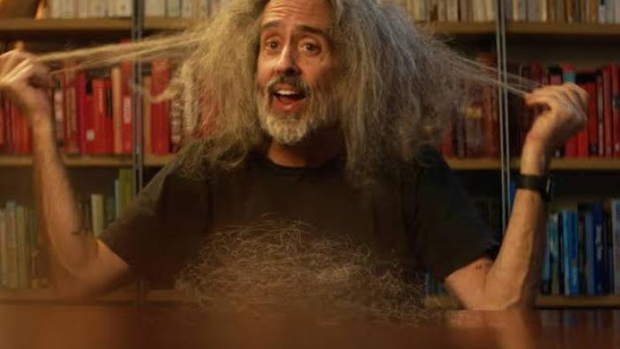 Andre is an Idiot: Man with wild hair pulling it outward, sitting in front of a colorful bookshelf.