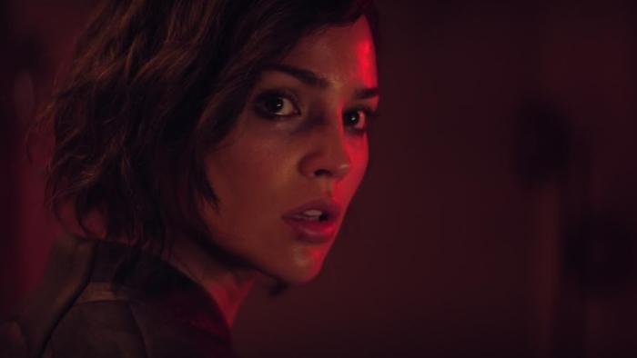 Ash Movie: Riya (Eiza González), face illuminated by a red glow, looks warily over her shoulder