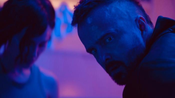Ash Movie: Brion (Aaron Paul, right), face illuminated by a blue glow, has a serious talk with Riya (in the background)