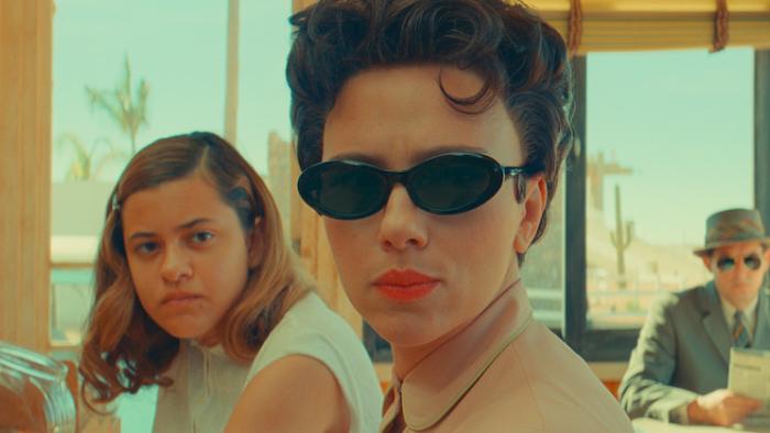 Asteroid City Movie: A woman in the 1950s wearing black sunglasses and red lipstick gives a hard look while sitting next to a teen girl