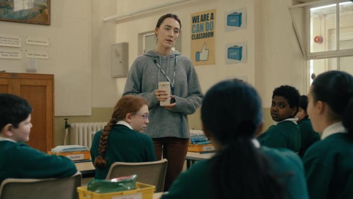 Bad Apples: White female teacher in a classroom, interacting with seated students wearing green uniforms