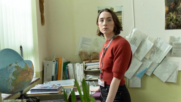 Bad Apples: White woman in school office looking despondent, standing near cluttered desk, globe, and papers on wall