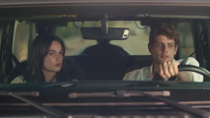 Bad Influence movie: White Spanish older teen girl with long brown hair left sits in front of car next to White Spanish young man in white shirt right