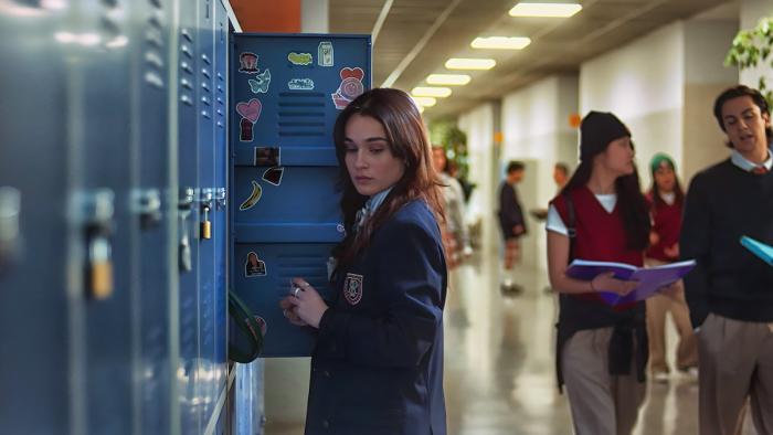 Bad Influence movie: White Spanish older teen girl in blue school jacket center stands next to blue lockers in school hallway, students to her right