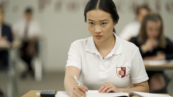 Bad Genius Movie: Lynn (Callina Liang) sits at a desk wearing a white uniform, taking a test, a defiant look in her eyes