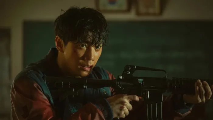Badland Hunters movie: Korean man with automatic rifle center stands in a classroom