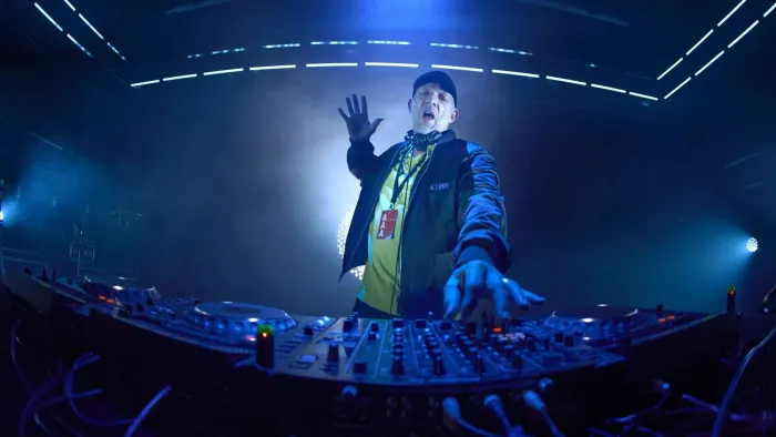 Banger movie: White French man in baseball hat, jacket, center, DJs and dances in front of turntables and equipment, dark lights all around him 
