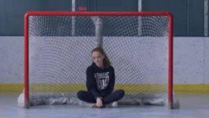 Before the Ice Melts movie: Girl sitting cross-legged in front of a red hockey goal net, wearing a navy sweatshirt.
